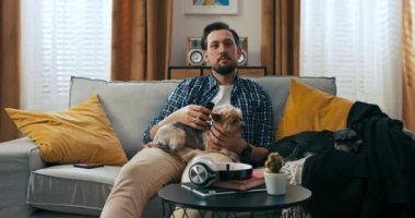 Mutlu sakallı adam kanepede oturmuş köpek dinleniyor uzaktan kumandayla kanalları değiştiriyor TV 'de ilginç haber filmleri seyrediyor..