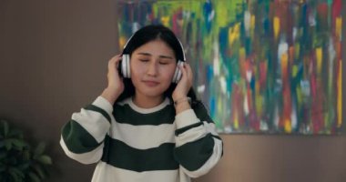 Genç bir kadın kablosuz kulaklıkla müzik dinlerken şarkının ritmine kafa sallıyor, kollarını göğsüne kavuşturuyor ve kameraya bakarken gülümsüyor..