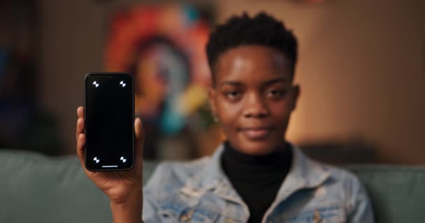 テクノロジー企業のためのデジタルマーケティング製品の専門家リビングルームのアフリカの女性は 拡張現実機能の仕事を革新的な機能を示す暗い画面で携帯電話を保持しています — ストック動画