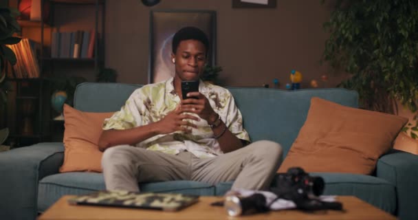 Afrikaanse Man Rust Zijn Bank Woonkamer Chatten Zijn Mobiele Telefoon — Stockvideo