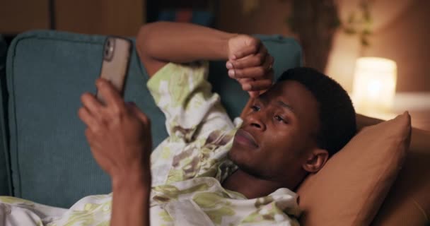 アフリカの男性は リラックスと接続を見つける 彼の携帯電話上の友人との活発なチャット会話に巻き込まれ リビングルームのソファに座っている — ストック動画