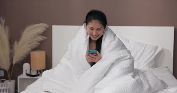 アジアの若い女性は 起きた後 電話でニュースをチェックします 世界のニュース 白いキルトで覆われた少女が快適なベッドに座る — ストック動画