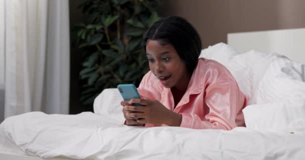 女性は手元に電話でベッドの上に快適に横たわっています ガールは オンラインストアを閲覧する笑顔 スマートフォン経由で友達とチャット — ストック動画