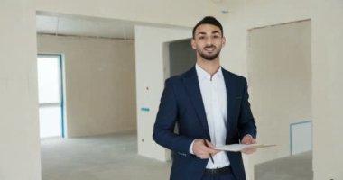 Orta Doğulu genç emlak yöneticisi müşterilerini karşılıyor. Yeni modern bina binasında müşterileri kameraya el sallayarak merhaba diyor..