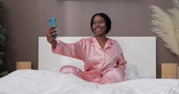 若いインフルエンサーが目覚めた後にベッドで自分自身の写真を撮ります ピンクのパジャマを着た女性は スマートフォンを手に持ち 自分撮りをする — ストック動画