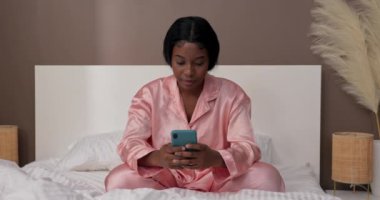 Gülümseyen mutlu kadın yatak odasındaki pijamalarıyla oturur ve elinde bir telefon tutar. Arkadaşına mesaj gönderiyor, komik resimlere bakıyor ve gülüyor..