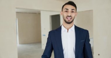 Orta Doğulu genç emlak yöneticisi müşterilerini karşılıyor. Yeni modern bina binasında müşterileri kameraya el sallayarak merhaba diyor..