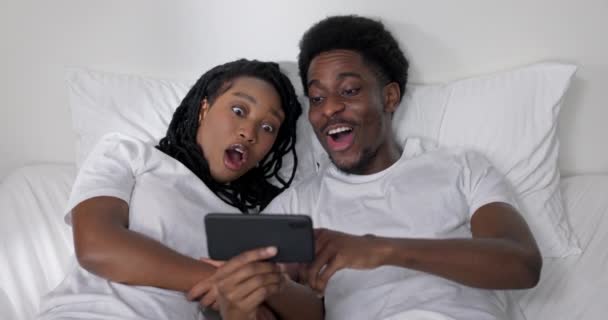 快乐快乐的美国黑人家庭坐在床上 带着手机微笑看着智能手机屏幕 和朋友们一起打视频电话 使用新的移动应用程序 在家里观看放松的视频 — 图库视频影像