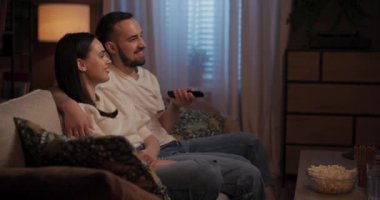 Oturma odasındaki koltukta oturan çift, uzaktan kumandalı bir adam patlamış mısır yerken televizyon kanallarını değiştiriyor. Programlar hakkında yorum yapar ve sıkıntı veya ilgi gösterirler..