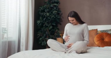 Kısa saçlı hamile bir kadın, karnına dokunduğunda neşe ve minnettarlık saçarak yatağında oturur. İçinde büyüyen küçük mucizenin beklentisiyle doludur. Kutsanmış Beklentiler konsepti.