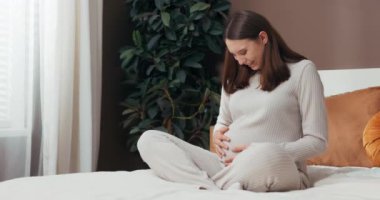 Neşe kavramı beklenirken mutlu gülümseyen, kısa saçlı hamile bir kadın yatağına oturur, sevgi dolu bir şekilde karnına dokunur ve ufaklıkların gelişini heyecanla bekler..