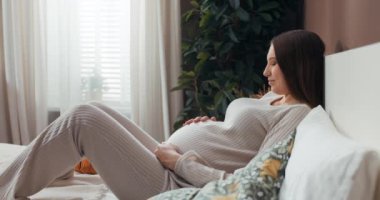 Hamile bir kadın rahat ve modaya uygun pijamalar içinde, eli karnında dinleniyor ve bebeğiyle bir anlık mutluluk yaşıyor..
