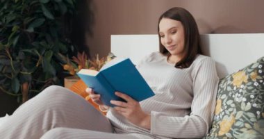 Gülümseyen hamile kadın rahat bir yatakta oturup kitap okuyor ve bebeği gibi kahkahalarla gülüyor..