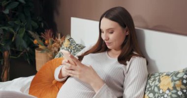 Hamile bir kadın yatağa uzanıyor, bebeğiyle konuşmak için yere koymadan önce bir cep telefonu tutuyor, şefkatle karnına dokunuyor..