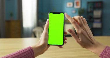 Oturma odasındaki kanepede rahatça oturan bir kadın. Elinde bir cep telefonu tutuyor ve ekranda canlı yeşil bir model görünüyor. Cihazı aktif olarak kullandığını gösteriyor..