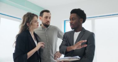 Bulaşıcı bir gülümseme ile Afrikalı emlakçı titizlikle detaylı zemin planı tutar, yol gösterici çiftler açık konsept yaşam alanına bakar, sonsuz olasılıkları vurgular..