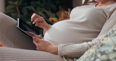 Yakın planda hamile bir kadın elinde tabletle fotoğraflara odaklanmış bir şekilde karnına dokunuyor. .