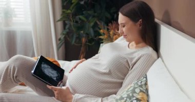 20 'li yaşlarının sonundan 30' lu yaşlarının başına kadar güzel hamile bir kadın şık ve rahat bir kıyafet giymiş bir yatakta oturuyor ve tablet üzerinde fotoğraf çeviriyor..