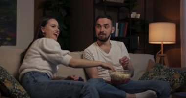 Oturma odası TV ekranından gelen tek ışık kaynağı genç kadın ve erkek kanepede yan yana oturur filme dalmış halde ara sıra birbirlerine patlamış mısır fırlatmak ve kontrolsüz bir şekilde kıkırdamak için dururlar..