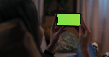 Uzun siyah saçlı, elinde yeşil ekranlı cep telefonu olan bir kadın yatay manzara pozisyonunda komik film videoları seyrediyor..