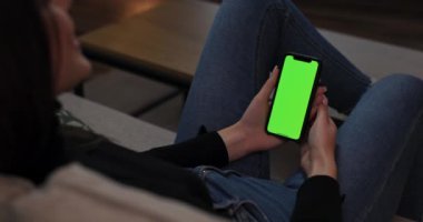 Kanepede oturmuş yeşil model ekranlı cep telefonuyla günlük kıyafetli esmer bir kadın görüntüsü. Bakış açısı.