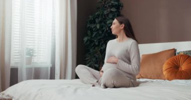 Yüzünde huzurlu bir gülümsemeyle, yatağında kısa saçlı hamile bir kadın, karnına nazikçe dokunurken, doğmamış bebeğiyle huzur ve bağ kuruyor. Dinginlik anı kavramı.