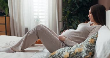 Şık pijamalı bir kadın olarak anneliğin güzelliği karnına hafifçe dokunur, sevgi ve beklentiyle parlar..