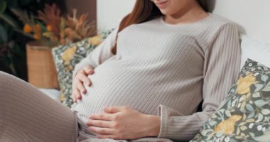 Sıcak pijamalarıyla, karnını nazikçe okşayan, küçük kızının gelmesini beklerken düşüncelere dalmış bir kadının yakın çekim görüntüsü..