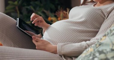 Yakın planda hamile bir kadın elinde tabletle fotoğraflara odaklanmış bir şekilde karnına dokunuyor. .