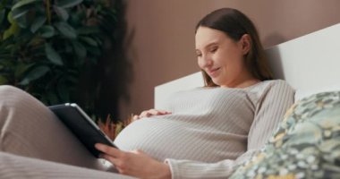 Modayı takip eden ve rahat giyimli hamile bir kadın elinde tabletle bir yatakta oturuyor ve yüzünde bir gülümsemeyle fotoğraflara bakıyor..