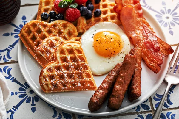 Breakfast table with waffles, fried egg, bacon and sausage served with fresh berries and maple syrup