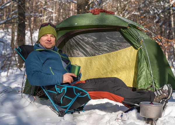 A man near the tent prepares to eat. Winds up the rope. Night in the winter forest. Love for nature. Eco tourism. Sun, forest, snow. Active lifestyle . Tent in the snow.