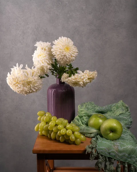 Bouquet of white dahlias in a lilac vase on a gray background, wooden table. Nearby lies a large bunch of green grapes, two green apples and a green handkerchief. Still life. Vitamins.Holiday.