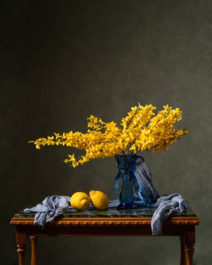 Mermer ahşap bir masanın üzerinde sarı çiçek dalları. Yakınlarda iki limon ve mavi bir mendil var. Koyu gri arka plan. bahar hala hayattır. Vitaminler. Yiyecekler. Diyet olsun. Sağlık.