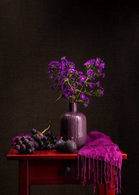 Bir buket leylak kasımpatı. Kırmızı ahşap bir masanın üzerinde mor bir vazoda küçük sonbahar çiçeklerinin çeşitli dalları. Yakınlarda büyük bir demet mavi üzüm ve iki olgun mavi incir var. Koyu pembe eşarp 