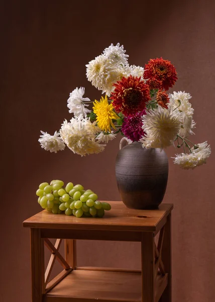 large bouquet of white, yellow, red, autumn flowers. Many asters in a gray vase on a wooden table, next to it lies a large bunch of green grapes. Orange background. Still life, postcard.