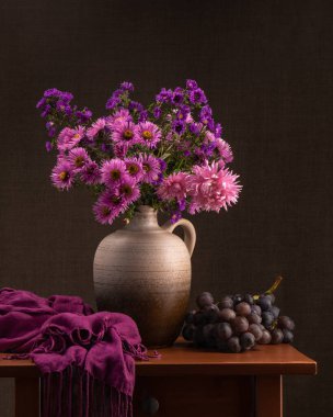 Gri kil vazoda bir sürü pembe, mor çiçek, tahta bir masada sürahi. Yakınlarda koyu gri bir şal ve büyük bir demet mavi üzüm bulunur. Gri arka plan. sonbahar buketi