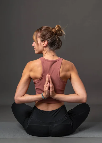 A young, athletic, attractive woman practices yoga. He sits with his back, arms crossed behind his back. The head is turned to the side. The hair is gathered in a bun. On a gray background
