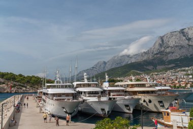 Hırvatistan 'ın Makarska kenti. Koyda demirli gemiler var. Toprak seti boyunca yürüyen insanlar. Güneşli bir gün. Dağ manzarası. Dağların üzerinde güzel bulutlar