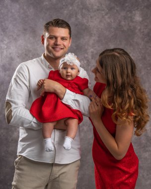 Beyaz gömlekli genç bir baba kırmızı elbiseli küçük bir kızı kollarında tutuyor. Kırmızı elbiseli anne nazikçe bebeğin yüzüne bakıyor.