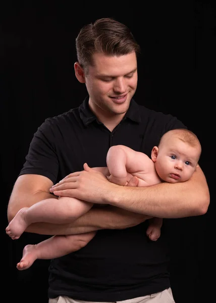 A young dad holds a newborn naked baby in his arms. Black background. Father\'s love.