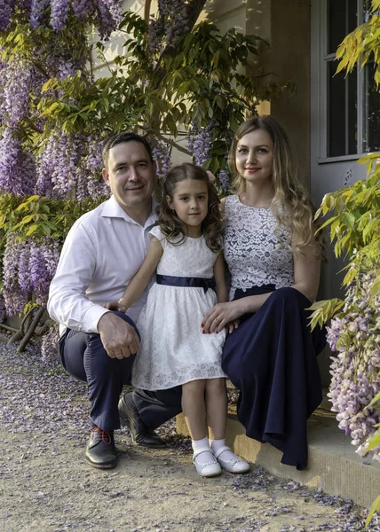 Family portrait. Mom, dad and daughter. Mom and girl are dressed in beautiful dresses, dad is wearing a white shirt. Floral background. Warm, close family relationships