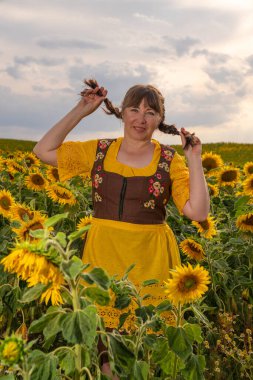 Bir kadın, sarı ayçiçekleriyle dolu bir tarlanın ortasında duruyor. Saçları örgülü, Alman ulusal kıyafetleri giymiş, sarı bluz, kahverengi elbise, sarı önlük. Batan güneşte.