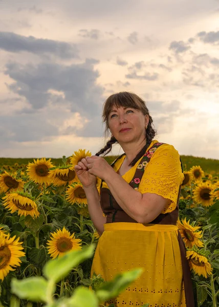 A woman is standing in the middle of a field with many yellow sunflowers. Hair braided, dressed in German national clothes, yellow blouse, brown sundress, yellow apron. In the setting sun