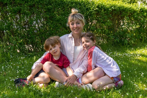 Modern mom sits on the grass and hugs cute little children,son and daughter. Cheerful family, joyful children. outdoor time with kids