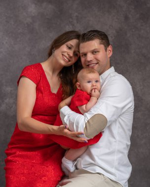 Anne ve baba küçük kızlarını kollarına alıp şefkatle birbirlerine bakıyorlar. Anne ve bebek kırmızı elbiseli, baba beyaz gömlekli. Stüdyo çekimi. Sevgi, aile