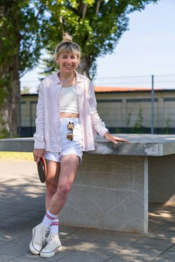 Modern bir kızın kısa şortlu portresi. Tenis masasının yanında duruyor, masa tenisi raketlerini elinde tutuyor. Neşeli bir şekilde gül. Açık havada..