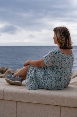 Orta yaşlı bir kadın sahilde oturur ve uzaktan bakar. Yaşamın, huzurun ve tatminin tadını çıkarıyor. Ufuktaki deniz ve dağlar.