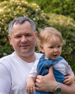 Bir baba ve küçük bir oğul portresi. Mutlu bir baba kollarında dalgalı, sarı saçlı küçük bir bebek tutuyor. Çocuk daha bir yaşında. Yakın plan. 