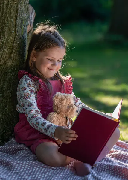 Parktaki büyük bir ağacın yanında küçük, sevimli bir kız oturuyor. Bir kitap okuyor, kollarında küçük bir oyuncak ayı oturuyor..